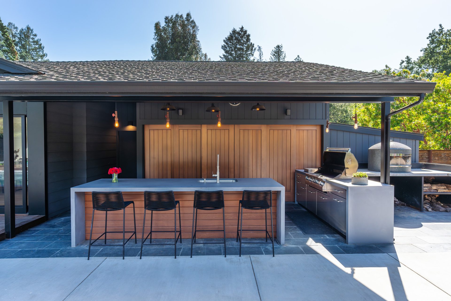 Modern outdoor kitchen with a concrete island bar, wood cabinetry, built-in grill, and a domed pizza oven