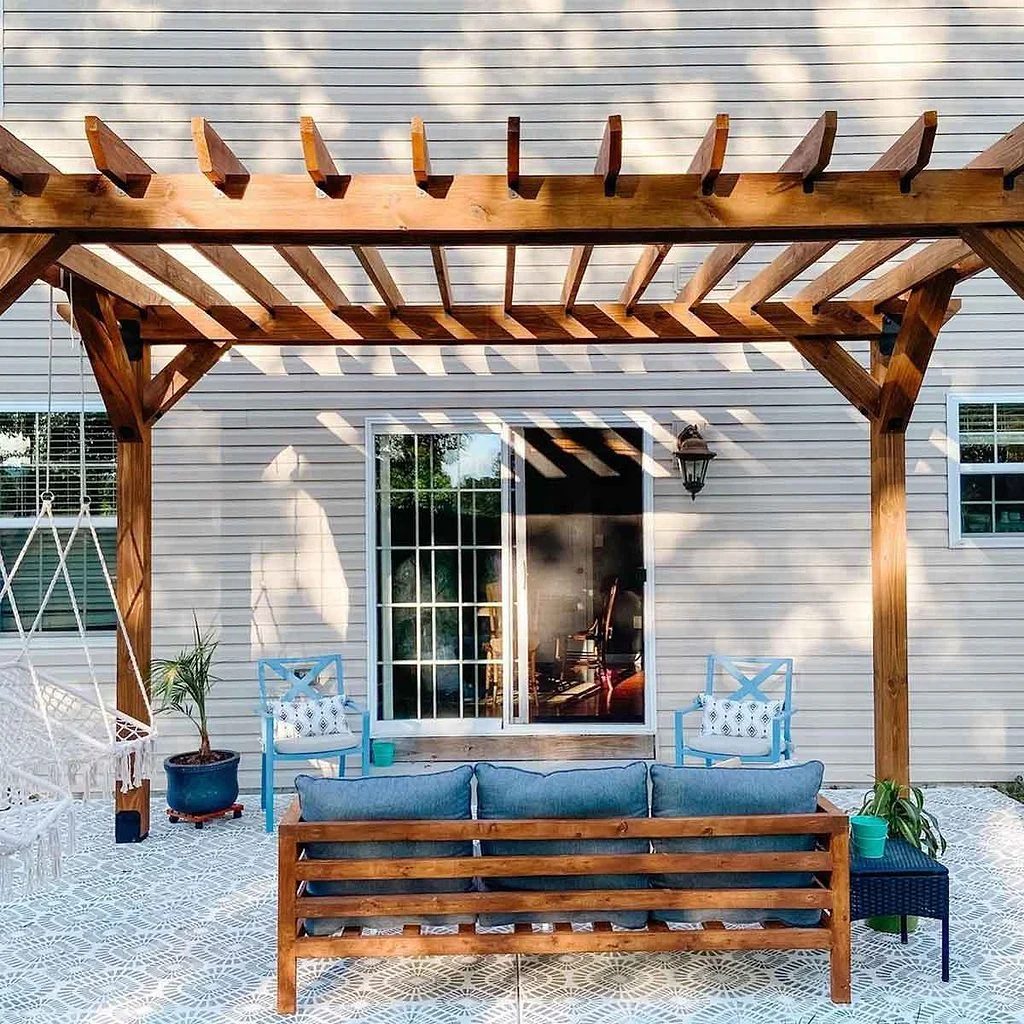 Wood pergola over a paver patio with a slatted sofa, hanging chair, and potted plants against a home