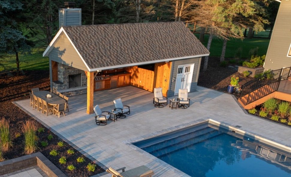 Poolside pavilion with an outdoor kitchen, stone fireplace, and patio seating beside an in-ground pool at golden hour