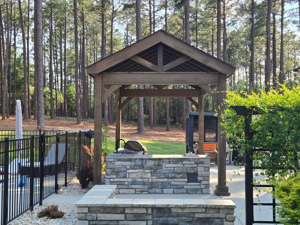Timber-frame pavilion with a shingled gable roof sheltering a stone outdoor kitchen beside a fenced pool