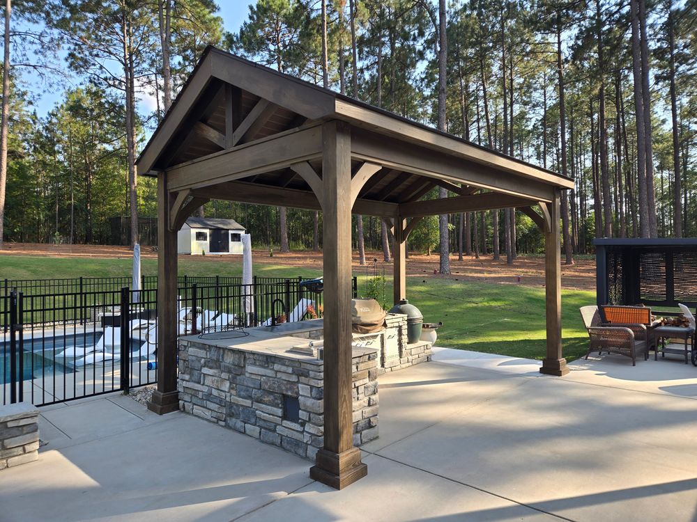 Wood pavilion and stone outdoor kitchen next to a pool, backed by tall Carolina pines at golden hour